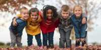Children stand arm in arm wearing coats in a fall scene.